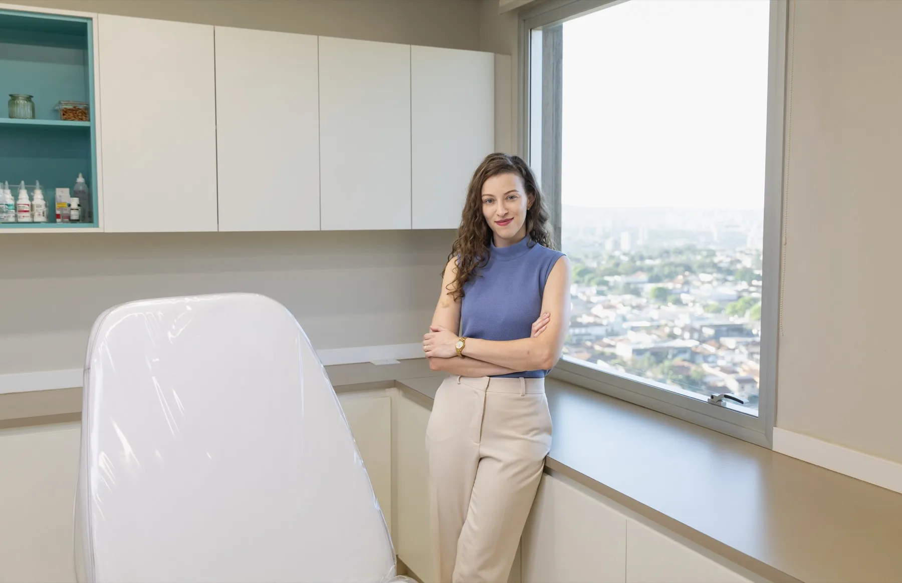 Dra. Natália Portelinha no consultório, ao lado da maca de exame e da janela com vista para Ribeirão Preto.